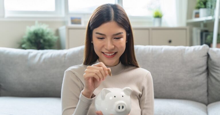 Young woman saving money monthly expenses putting coin in to piggy bank on the table.